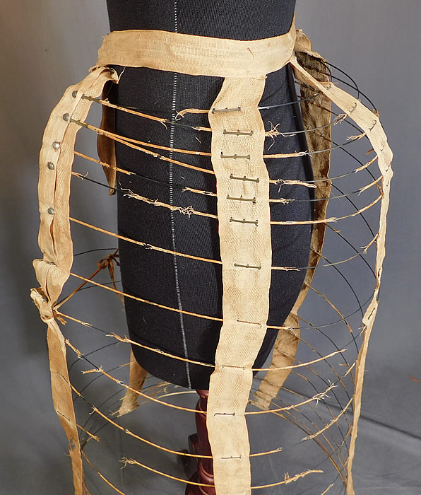 Victorian Bustle Back Wire Hoop Crinoline Cage Petticoat Skirt Support
This wonderful womans collapsible cage petticoat has a protruding bustle hump on back and an adjustable waistband strap with buckle clasp closure on the front. This unique undergarment is a type of framework that would have been worn under a bustle dress to expand the fullness and support the back drape of the fanned train skirt.