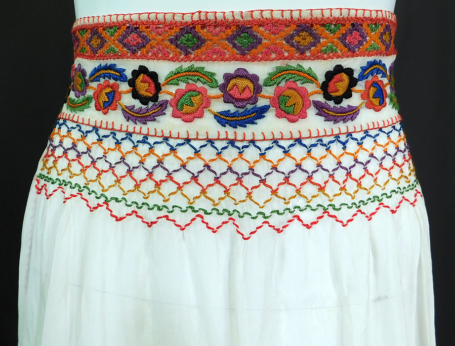 Vintage White Cotton Colorful Floral Embroidered Smocking Boho Peasant Half Apron Skirt
This vintage white cotton colorful floral embroidered smocking boho peasant half apron skirt dates from the 1920s. It is made of a sheer white cotton muslin fabric, with a colorful raised padded satin stitch hand embroidery work floral vine leaf design, multi colored diamond pattern lace trim, smocking stitching and red whip stitch edging. 