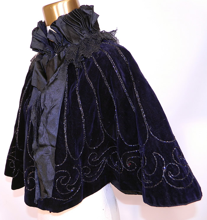 Victorian Black Velvet Jet Beaded Evening Opera Cloak Cape Capelet. This elegant evening opera cloak cape capelet has a pleated black silk ruffle stand up collar with a black lace ruffle trim, black silk ribbon tie drape down the front, hook closures and is fully lined. The cape measures 17 inches long, with a 112 inch bottom circumference and 15 inch neck. 